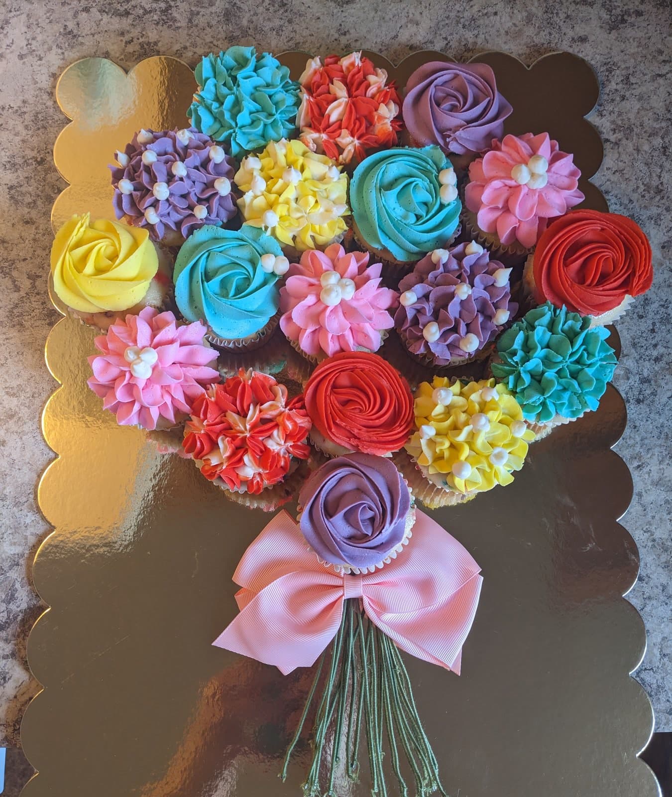 A bouquet of cupcakes arranged as a posy of piped buttercream flowers in pink, purple, yellow, blue and red, gathered with a pink ribbon bow on a gold scalloped board