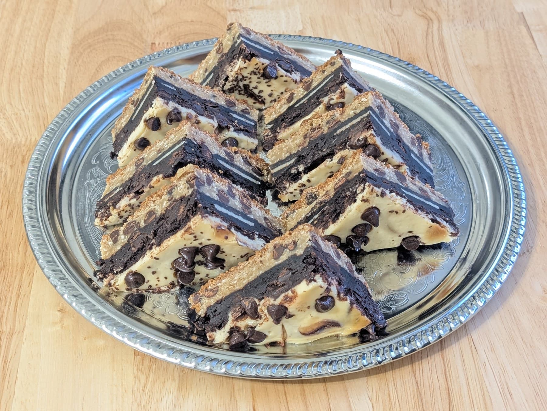 Triangular layered dessert bars — pale cookie dough studded with chocolate chips over dark cookie and brownie layers — arranged in rows on a silver platter