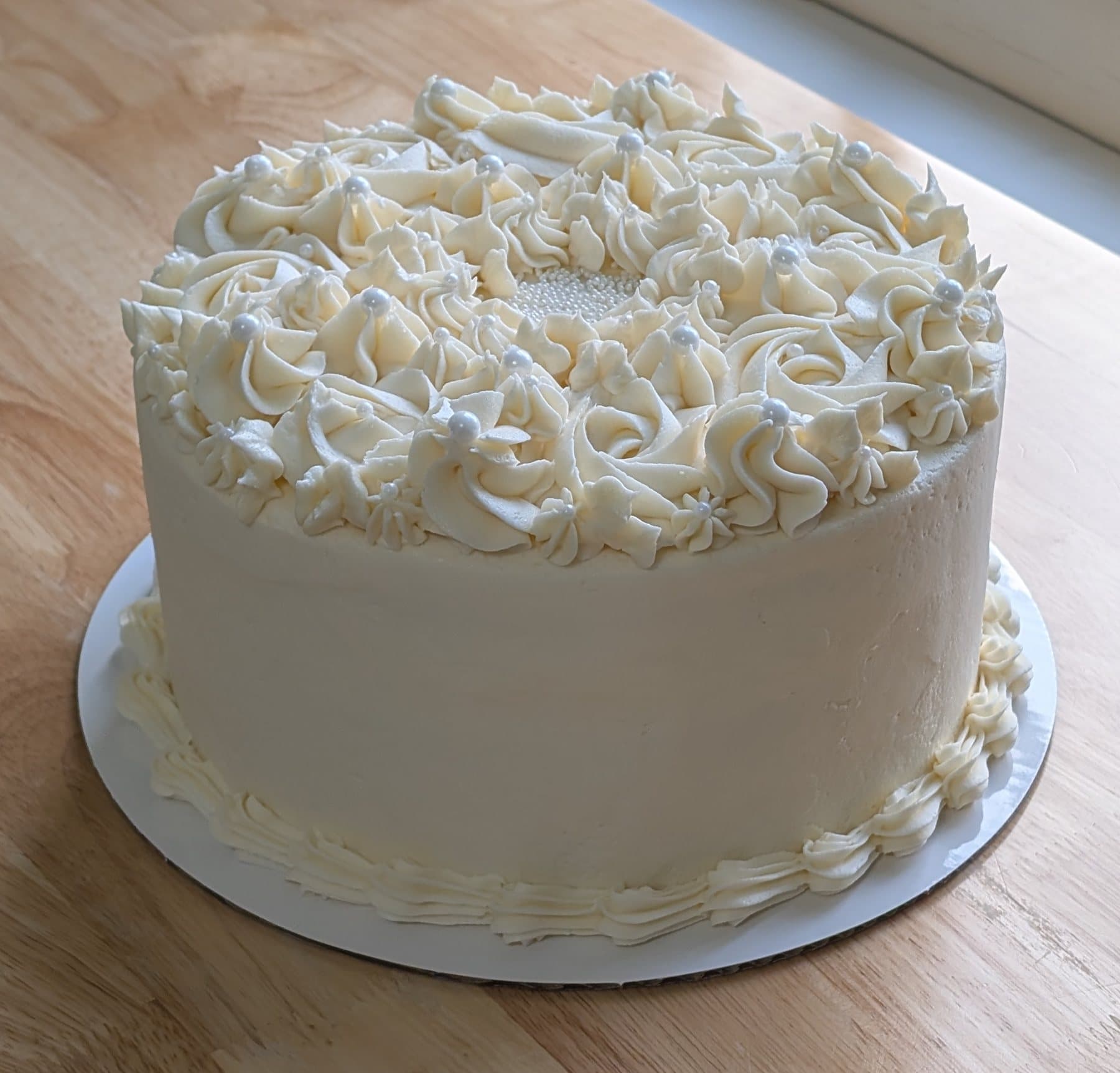 A round ivory cake in smooth buttercream, its top covered edge to edge with piped roses, rosettes and small star flowers studded with white pearls, on a white board on a wooden table