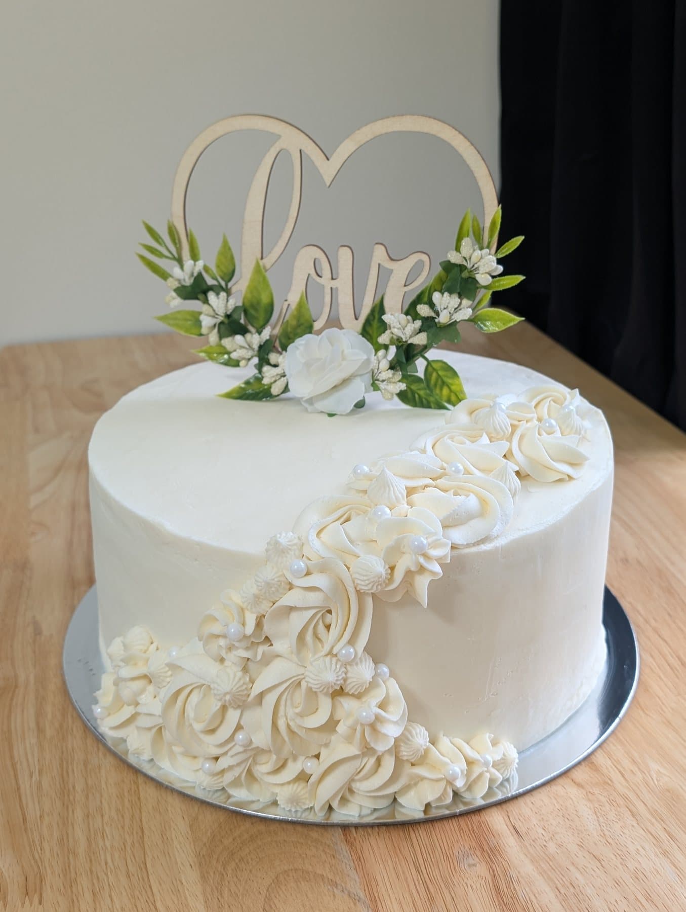 A round ivory cake in smooth buttercream with a cascade of piped rosettes and small flowers with white pearl accents down one side, topped with a pale natural wood heart topper reading Love, dressed with greenery and a white rose