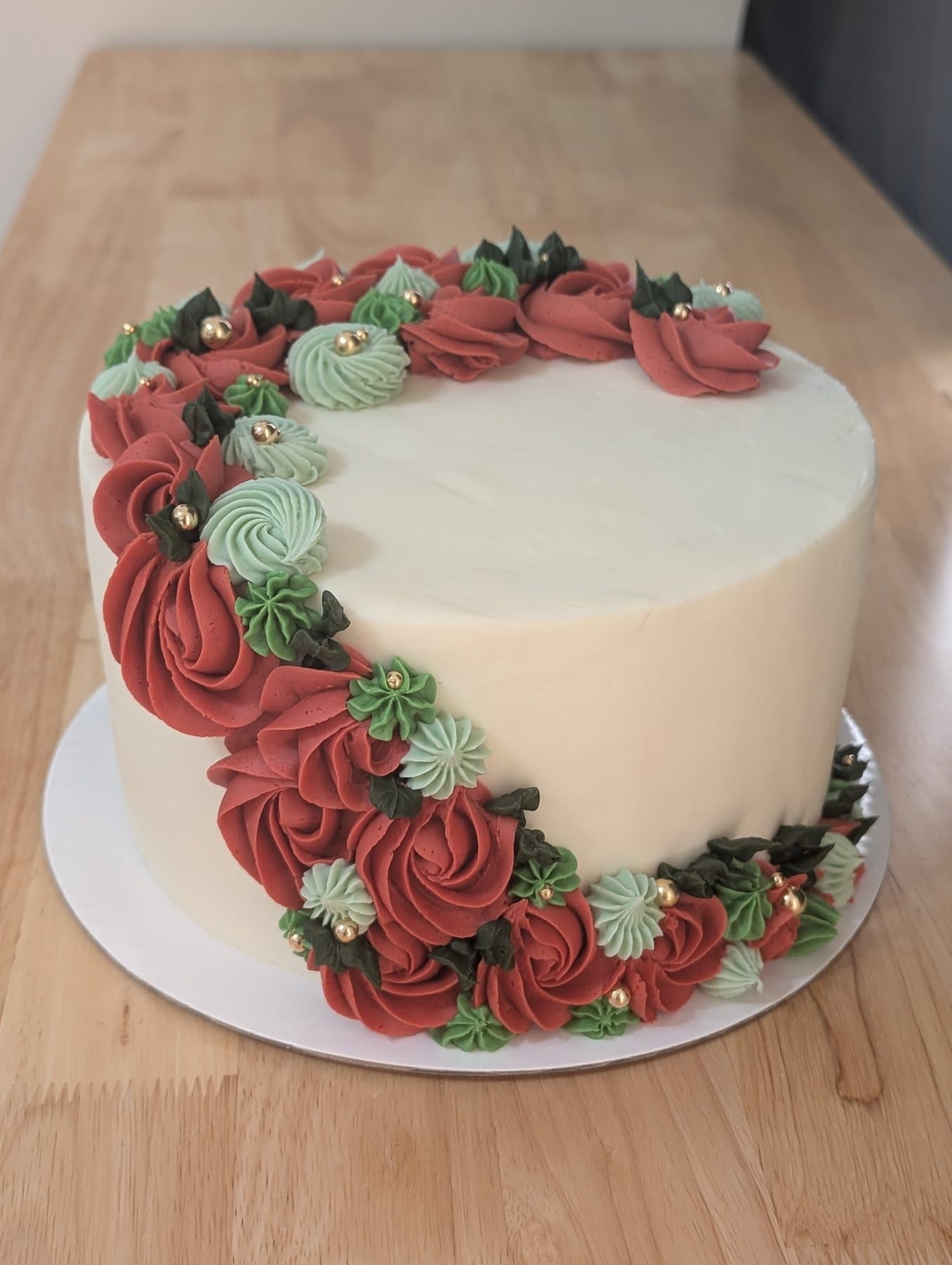 A round cake in ivory buttercream with a cascade of deep red piped roses and green piped leaves down one side