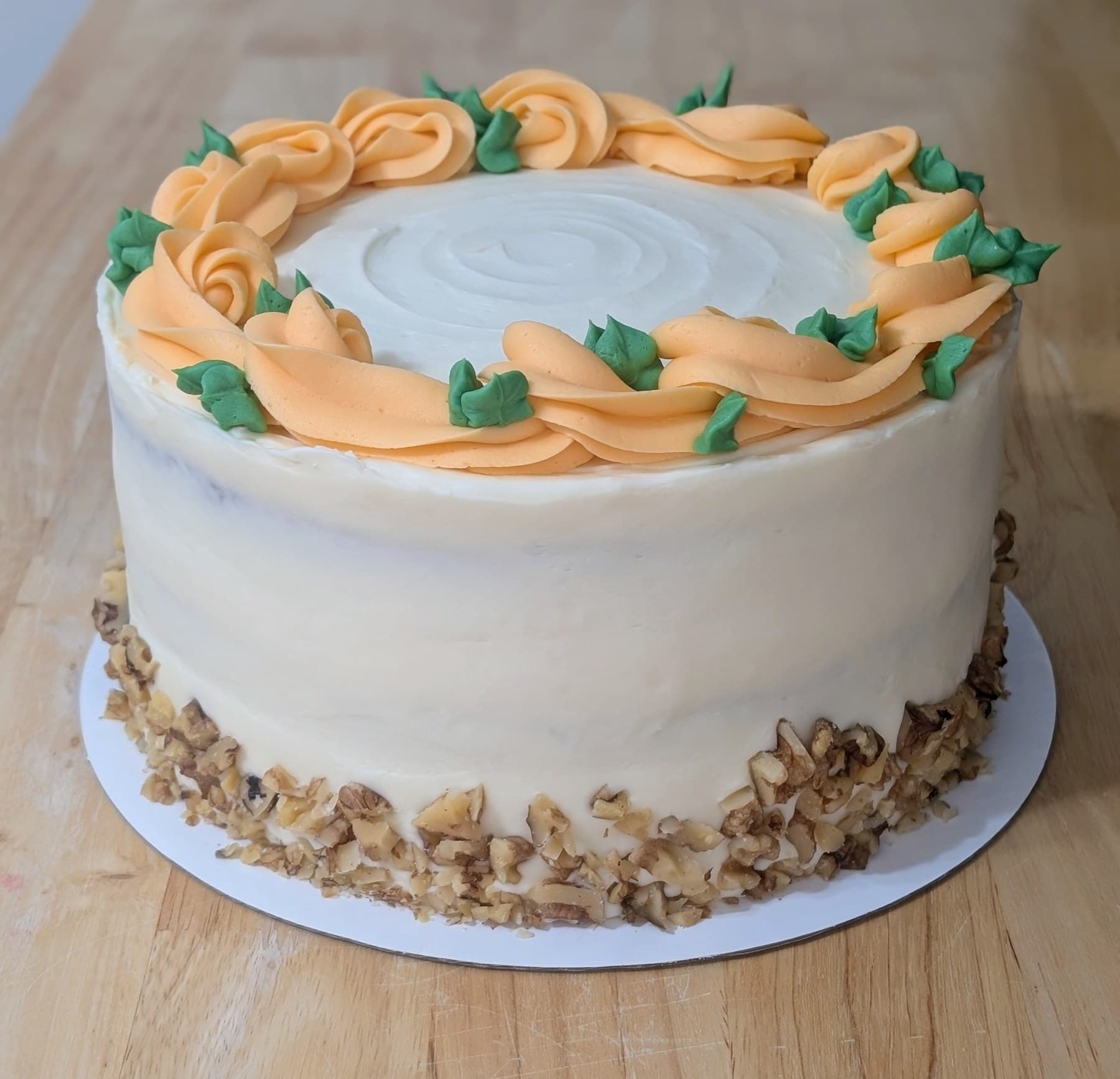 A round cake in smooth cream frosting, ringed on top with piped orange rosettes and green leaves, its base bordered with chopped nuts