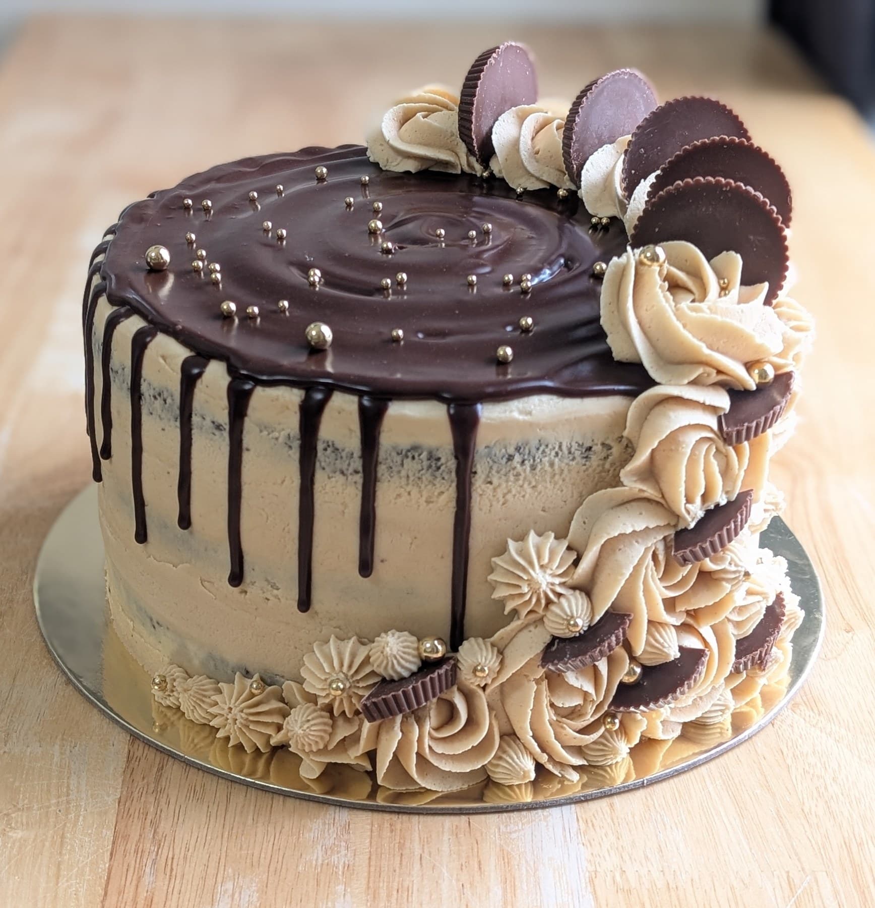 A cake in tan buttercream with a glossy dark chocolate top scattered with gold pearls, a cascade of piped rosettes and halved peanut butter cups down one side