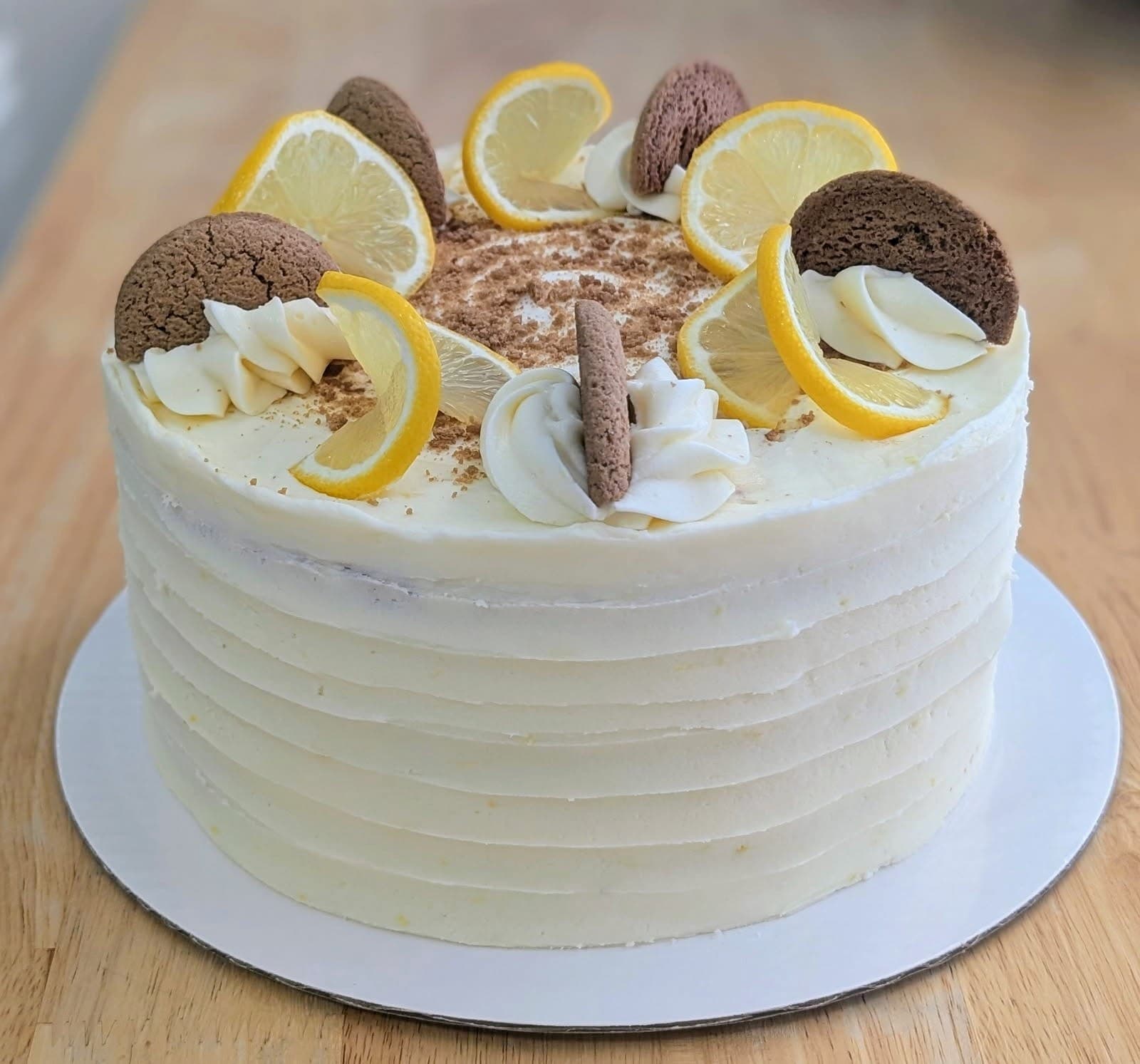 A round cake in pale lemon buttercream with combed sides, its top ringed with lemon slices, gingersnap cookies and piped swirls around a centre dusted with cookie crumbs