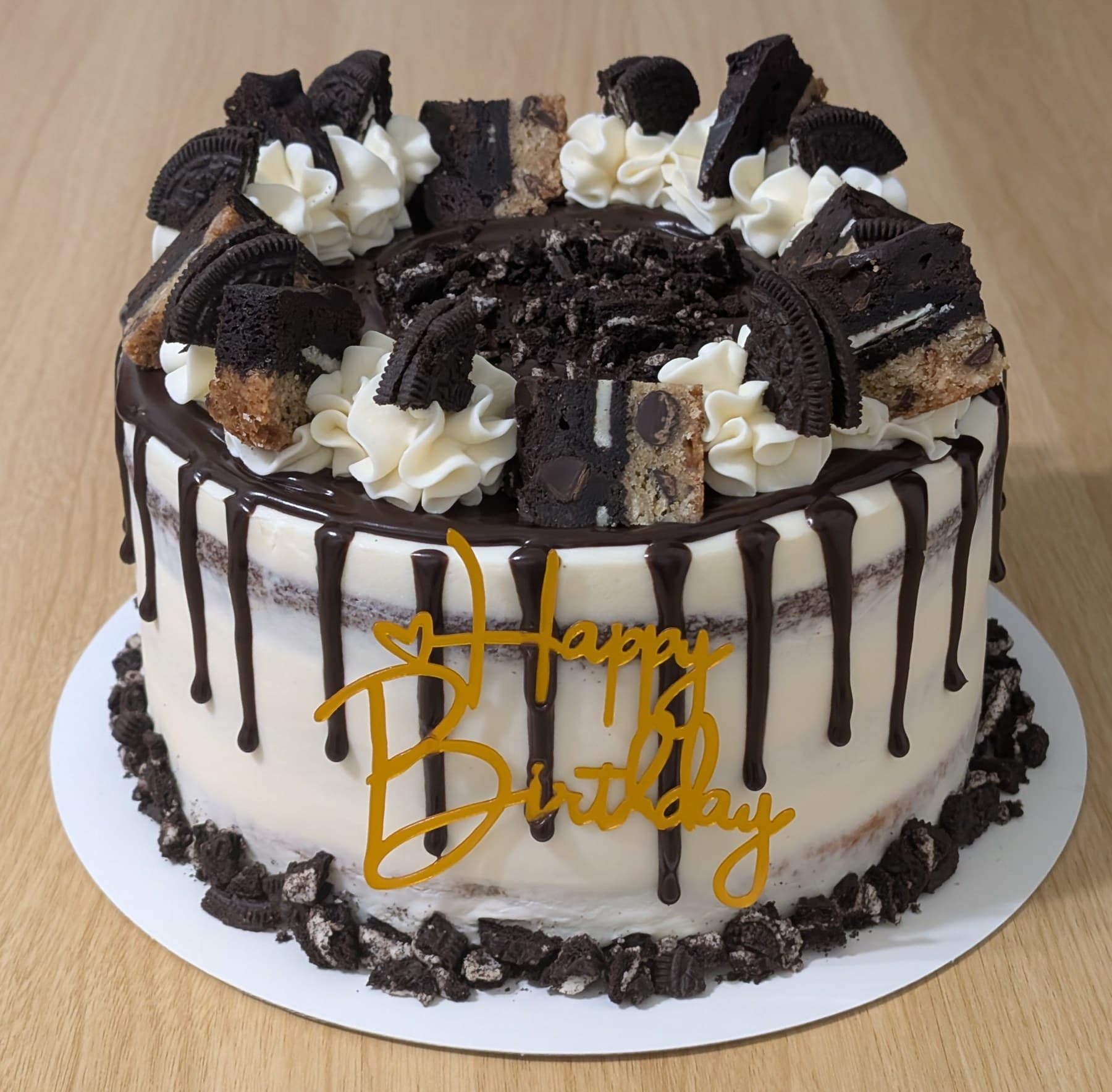 A semi-naked cake with a dark chocolate drip, its top piled with sandwich cookies, brownie and cookie pieces between piped ivory swirls, with a gold Happy Birthday topper