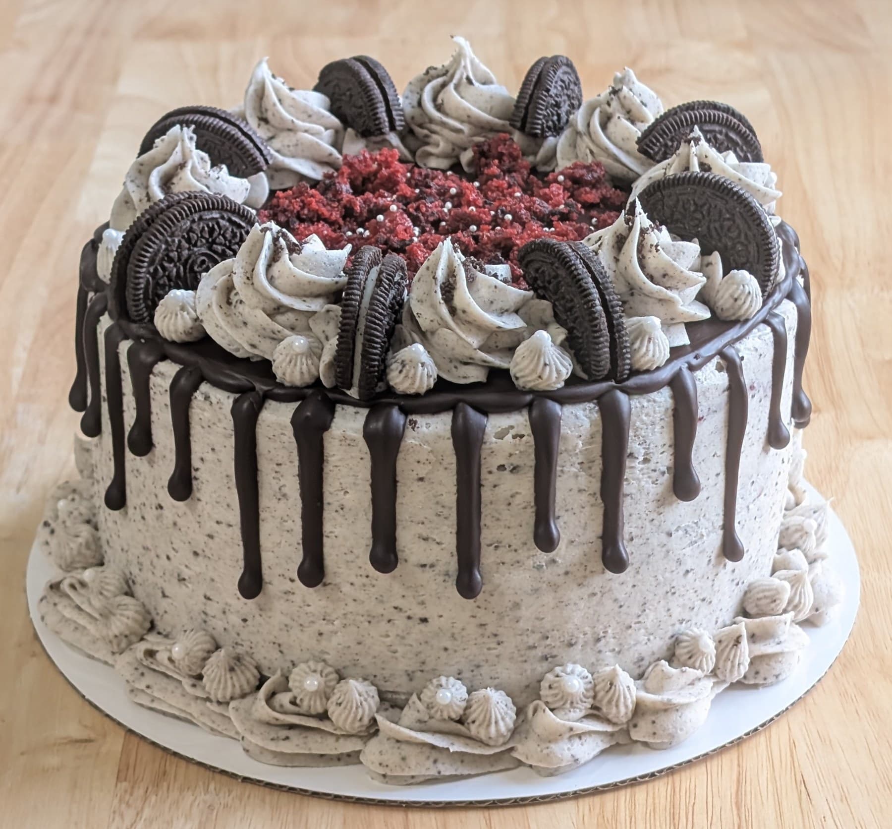 A cookies-and-cream frosted cake with a dark chocolate drip, its top ringed with sandwich cookies and piped swirls around a centre of red velvet crumbs