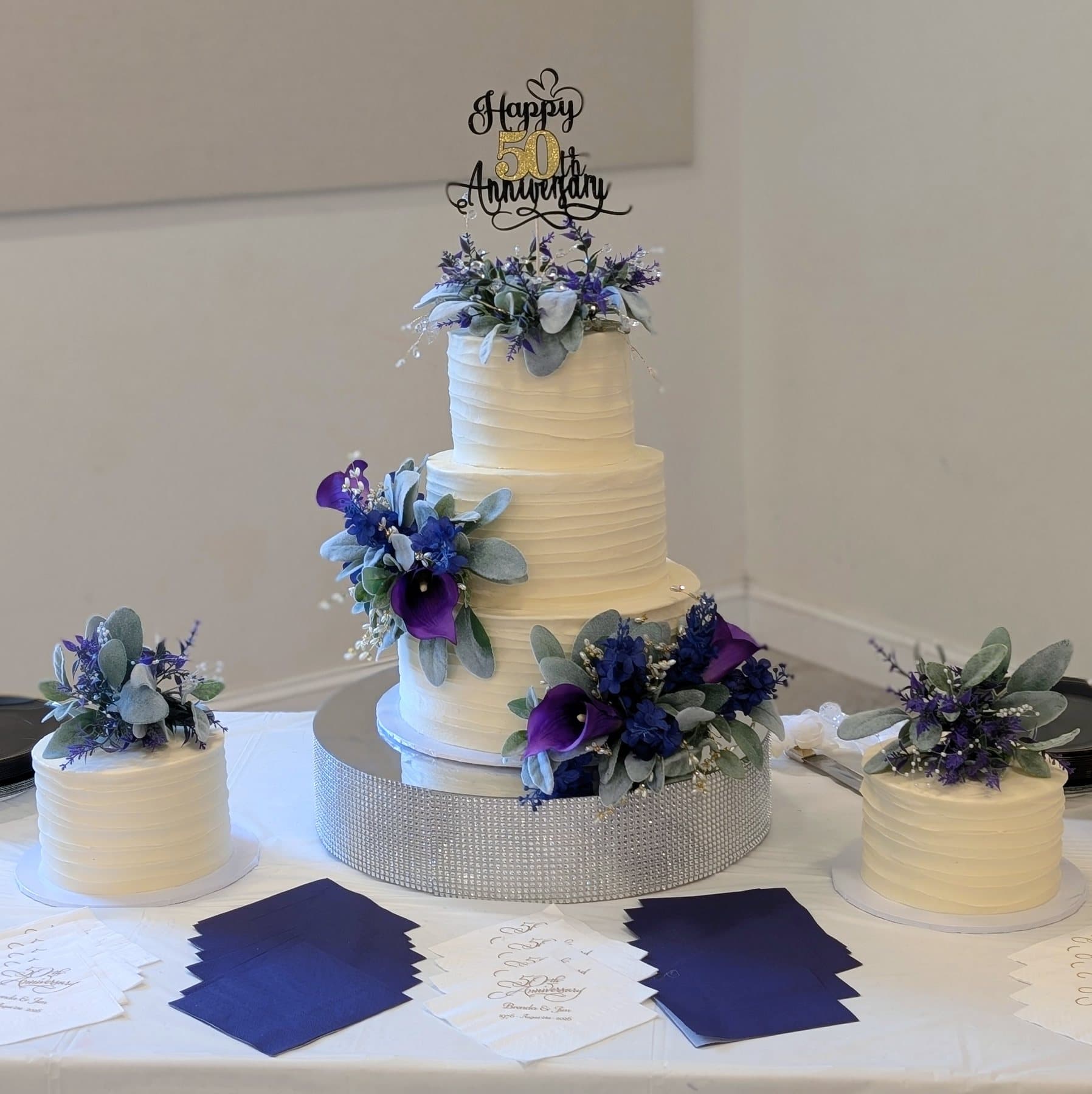 A three-tier textured buttercream cake dressed with purple calla lilies and blue florals, flanked by two matching single-tier cakes on the same table