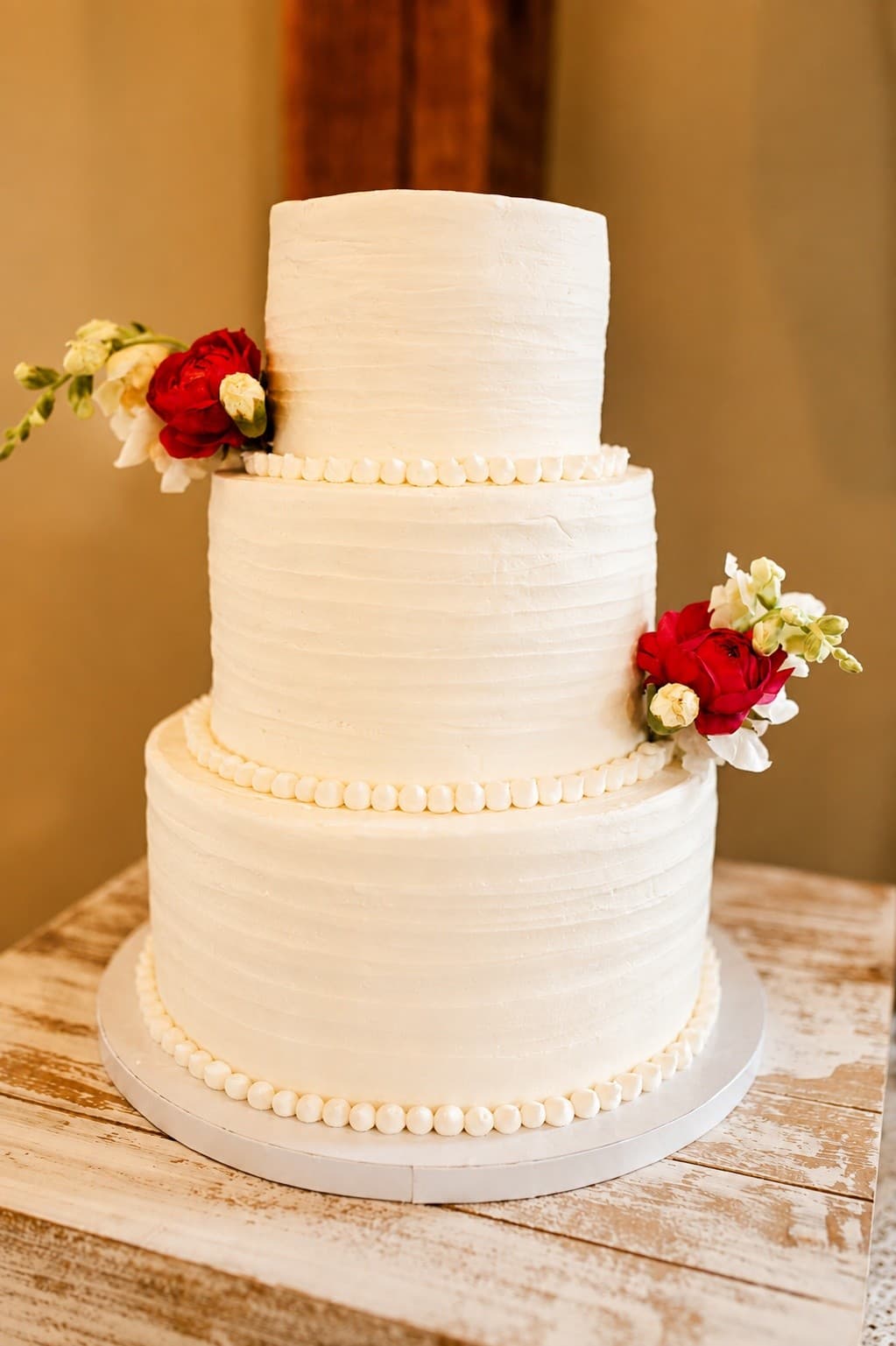A three-tier white wedding cake in textured buttercream with piped pearl borders between the tiers and clusters of deep red roses and cream blooms